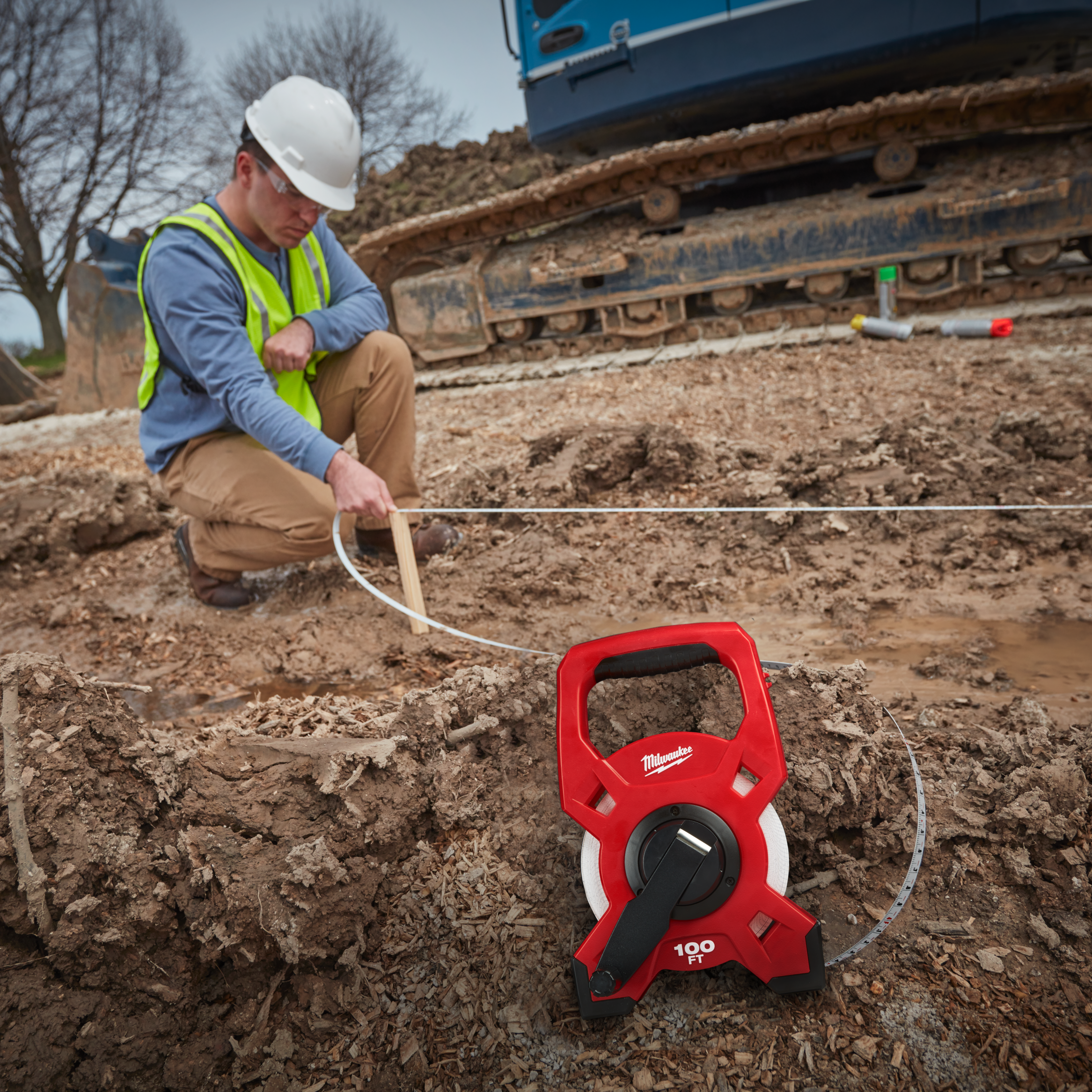 Person using a Milwaukee measuring tape in an excavation site with construction equipment in the background.