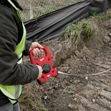 A person wearing a high-visibility safety vest holds a red Milwaukee tape measure against a dirt and gravel outdoor work site.