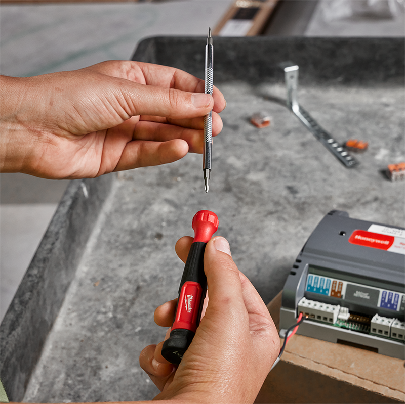 Person holding a screwdriver with a red handle and reversable bit holder, with an electrical box in the background