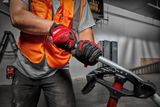 A worker wearing an orange vest and red-and-black Milwaukee gloves uses a tool in a workshop setting.