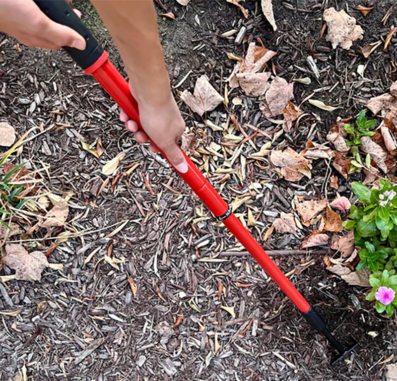 Person using a red garden tool on mulched ground with plants and flowers.