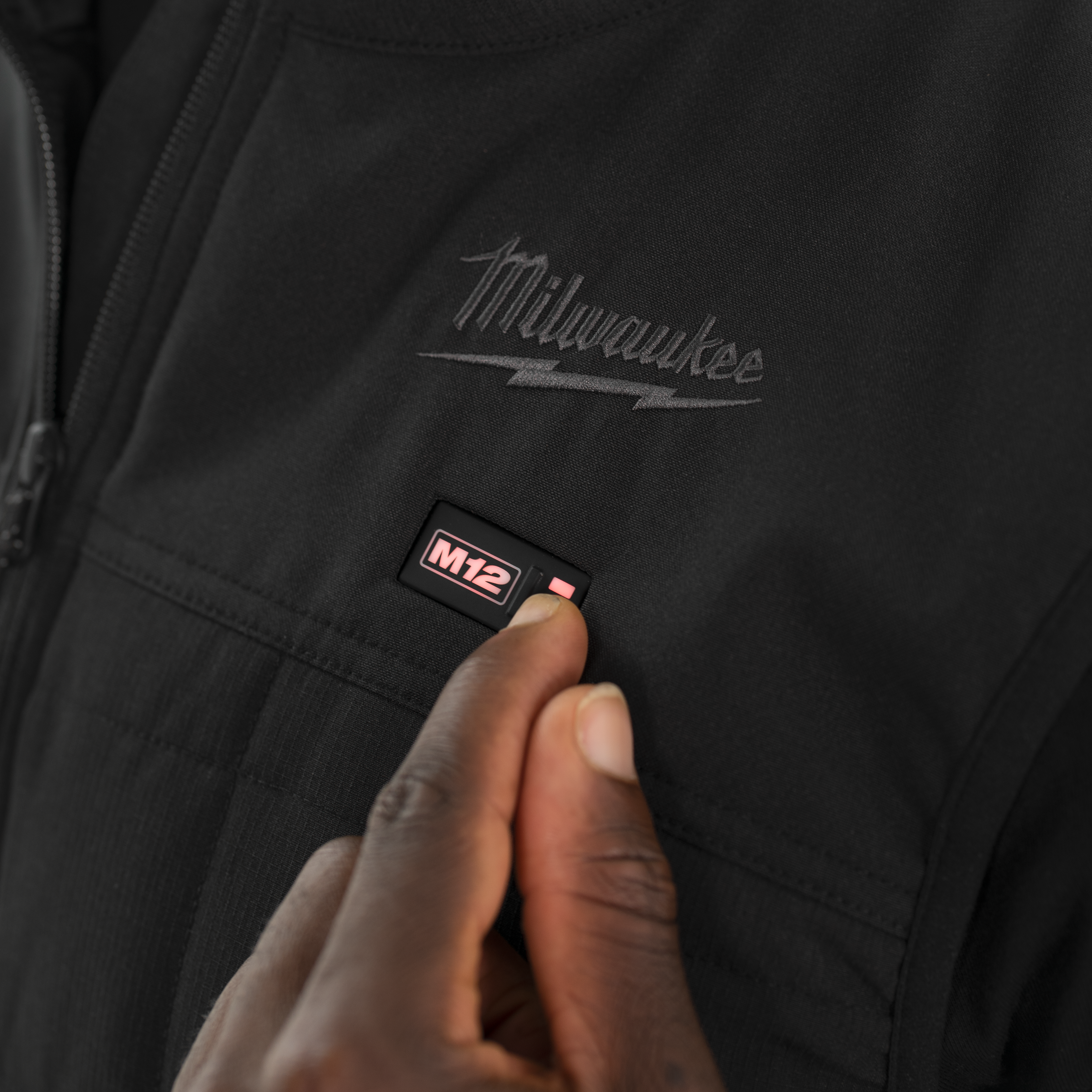 Black jacket with Milwaukee logo and M12 label, hand touching the jacket.